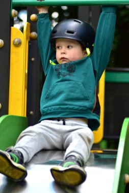 Çocuk parkta kaymaya hazır, kasklı ve yeşil kapüşonlu bir çocuk. Hareketli çekim odak, güç ve neşeli beklenti, güvenli ve enerjik açık hava oyunu sırasında..