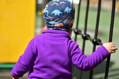 Arka planda, mor ceket ve dinozor şapkası giyen, oyun alanının iplerini keşfeden genç bir kız var. Fotoğraf, eğlence ve güvenli doğa aktiviteleri sırasında merak, bağımsızlık ve hareketi sembolize eder..