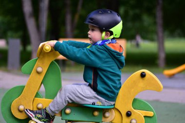 Çocuk yeşil ve sarı yaylı rockçı, oyun alanında motosiklet şeklinde, başlık ve kapüşonlu. Odaklanmış ifade aktif açık hava oyununun dengesini, eğlencesini ve neşesini gösterir.