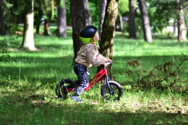 Yeşil parktaki ağaçların arasında kırmızı dengeli bisiklete binen kasklı küçük çocuk. Yan görünüm, açık hava macerasının odak, merak ve neşesini, aktif çocukluk yaşam tarzı ve doğa bağlantısının altını çizerek yakalar