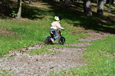 Yeşil parkta dengeli bisiklete binen kasklı küçük çocuk. Arka plan, çocuğun doğayı özgüvenle keşfettiğini, güneşli bir yaz gününde fiziksel aktiviteyi, özgürlüğü ve erken bisiklet becerilerini teşvik ettiğini gösteriyor..