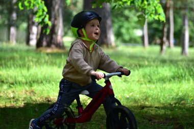 Yeşil parkta kırmızı dengeli bisiklete binen mutlu çocuk büyük bir gülümsemeyle yukarı bakıyor. Sahne heyecan, neşe ve kaygısız çocukluk keşiflerini yakalar, aktif ve sağlıklı oyunları teşvik eder..