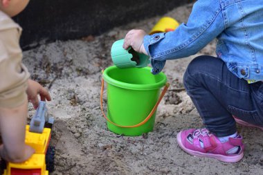 Yeşil kova ve oyuncak kamyonla kum havuzunda oynayan iki çocuk. Güneşli bir günde çocukların ellerine ve renkli oyuncaklara yakın çekim, açık hava eğlencesi, yaratıcılık ve çocukluk gelişimi..