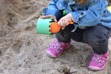 Kot ceketli küçük bir kız ve kum havuzunda oynayan pembe ayakkabılar, turuncu bir kürekle yeşil bir kürek içine kum topluyor. Açık hava etkinliği yaratıcılığı, motor becerileri ve yaratıcı oyunu teşvik eder..