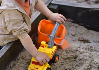 Sarı oyuncak kazıcısı ve turuncu kovayla kum havuzunda oynayan bir çocuk. Eller ve oyuncakların yakın plan görüntüsü, açık havada kumsalda oyun etkinliği, yaratıcılığı ve becerileri destekliyor..