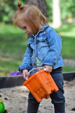 Kum havuzunda elinde turuncu kovayla duran kot ceketli, kızıl saçlı küçük kız. Sahne, doğada çocukluk eğlencesi, merak ve yaratıcı oyun zamanını tasvir eder, yaratıcı keşifleri teşvik eder.