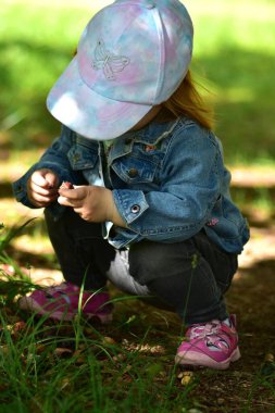 Pastel kelebek şapkası takan, kot ceket giyen, çimlere çömelmiş, ellerindeki küçük nesneleri keşfeden küçük bir kız. Sahne, merakı, duyusal öğrenmeyi ve sakin bir çocukluk anını ön plana çıkarır..