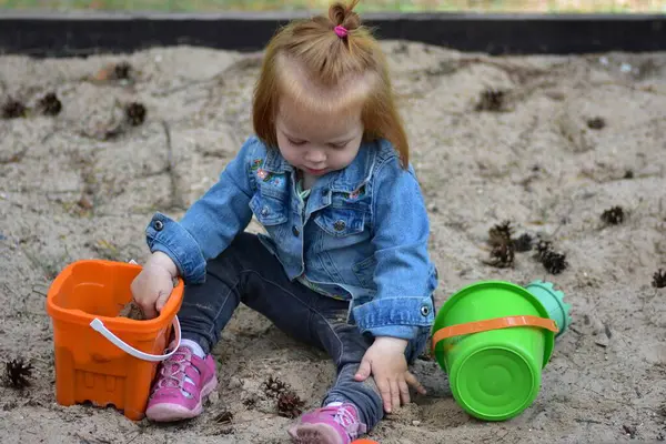 Turuncu kova ve yeşil kovayla kum havuzunda oturan küçük kız. Kum ve çam kozalaklarıyla oynar, doğada yaratıcı doğa eğlencesi, duyusal oyun ve erken çocukluk gelişimini anlatır.