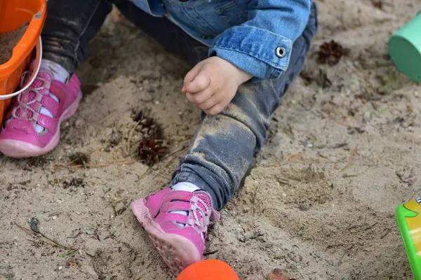 Kum havuzunda oynarken küçük kızın kumlu bacakları ve pembe ayakkabılarının yakın çekimi. Ayrıntılı görünüm dokunsal deneyimi, çocukluk karmaşasını ve duyusal keşfi, açık hava eğlencesini yakalar..