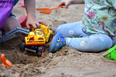 Sarı oyuncak kazıcı ve turuncu kürekle kum havuzunda oynayan iki çocuğun yakın çekimi. Sahne, takım çalışmasının ve yaratıcı açık hava oyununun bir anını yakalıyor..