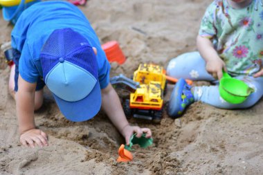 Erkek ve kız, inşaat oyuncakları ve kovalarla kum havuzunda yan yana oynuyorlar. Kız kumu yeşil bir konteynıra doldururken, mavi şapkalı çocuk kazıyor ve kooperatif oyun oynuyor..