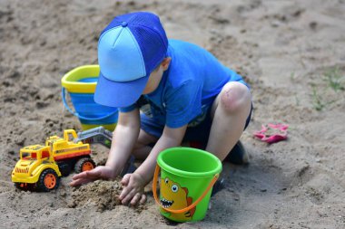 Mavi şapkalı ve gömlekli küçük çocuk kum havuzunu kazıyor oyuncak kamyonlar ve kovalarla, parlak bir yaz günü kumdaki ellerle oyuna odaklanıyor..