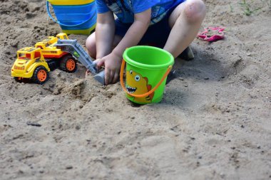 Güneşli bir günde kum havuzunda sarı bir oyuncak kamyonla oynayan ve etrafı kuru kum ve çimlerle çevrili iki kova çocuk, erken yaşta oyun oynarken yakalandı..