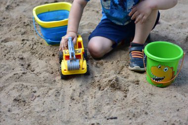 Güneşli bir günde, kum havuzunda sarı bir oyuncak kamyon ve iki kova ile oynayan anonim çocuk, erken çocukluk aktiviteleri sırasında yakalandı..
