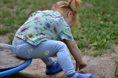 Renkli elbiseli genç bir kız oyun parkının yanındaki çamurla oynamak için eğiliyor. Erken çocukluk keşiflerinin sıradan bir açık hava sahnesinde yere odaklanıyor..
