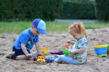 Mutlu çocuk ve kız bir kum havuzunda oyuncak kamyonlar ve kürekler ile birlikte oynuyorlar, hem gülüyorlar hem de kumda kazıyorlar. Çocukluk arkadaşlığının ve açık hava eğlencesinin neşeli bir sahnesi.