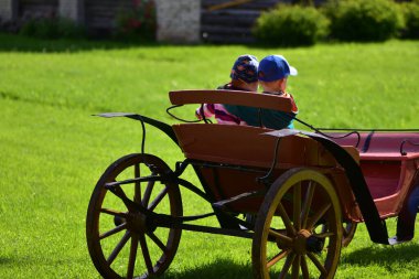 Güneşli bir çim tarlasında eski bir ahşap arabada şapkalı iki çocuk yan yana oturuyor. Arkadaşlık, çocukluk bağı ve doğal ortamda yakalanan klasik kırsal cazibe..
