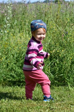 Renkli kıyafetlerle yeşil bir tarlada yürüyen neşeli küçük kız kameraya doğru gülümsüyor. Açık havada yaz eğlencesi ve tasasız çocukluk anları.