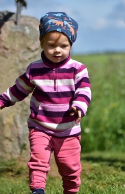 Güneşli bir günde arka planda çimen tarlası olan, üzerinde çizgili yün ve mavi bir şapka olan, dışarıda yürüyen bir kızın yakın plan portresi. Doğal çocukluk anı..