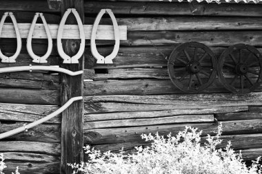 Geleneksel ahşap ahır duvarının siyah beyaz fotoğrafı. Eski araba tekerlekleri ve at boyunduruğu asılı. Eski kırsal mimari ve tarım ekipmanları sergileniyor.