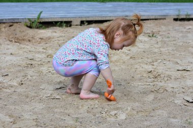 Küçük kız elinde turuncu plastik bir kürekle kumlu zeminde duruyor. Doğal bir arka bahçede merak ve hareketlerle dolu neşeli bir oyun zamanı yakalar..