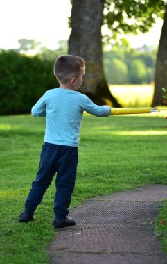 Açık mavi tişörtlü çocuk sarı su tabancası tutuyor, yaz akşamı çimlerin üzerinde duruyor. Ağaçlar ve tarlalar yumuşak arka plan ışığında. Dışarıda huzurlu bir çocuk anı..