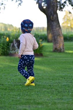 Küçük kız yeşil çimlerde yürüyor dinozor şapkası ve çiçekli pantolon giyiyor. Yumuşak akşam ışıkları ve arka planda ağaçlarla huzurlu bir kırsal alan..