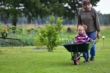 Gülümseyen yaşlı bir kadın yeşil bir el arabasıyla kırsal alanda neşeli kızıl saçlı bir çocuğu itiyor. Mutlu kırsal yaşam tarzı ve çoklu kuşak anları.