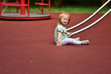 Kızıl saçlı çocuk kırmızı lastik oyun parkı yüzeyinde kaydırağın dibinde otururken gülümsüyor. Arkasından kırmızı bir atlıkarınca görünüyor, canlı bir sahne ekliyor..