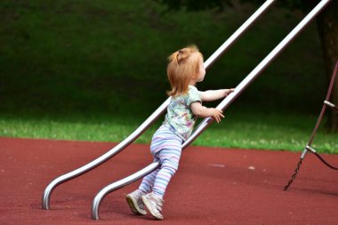 Kızıl saçlı çocuk halka açık bir oyun parkında parlak bir metal kaydırağına tırmanıyor. Çocuk odaklanmış görünüyor, yumuşak kırmızı kauçuk bir yüzey üzerinde kararlılıkla parmaklıkları tutuyor..