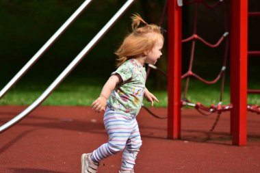 Kauçuk oyun alanı yüzeyinde yeşil çimlerle çevrili kızıl saçlı çocuk neşeyle koşuyor. Çocuk enerjik ve güneşli bir günde renkli oyun kıyafetleri giyiyor..