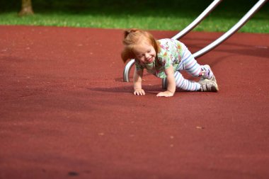 Kauçuk bir oyun parkında kaydırağın yanına düştükten sonra sevinçle gülen kızıl saçlı çocuk. Çizgili tayt ve renkli bir gömlekle, neşeli bir ifadeyle aktif oyundan hoşlanıyor..