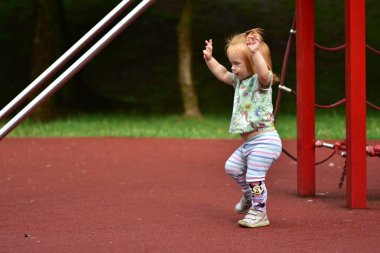 Küçük kız oyun parkında neşeyle oynuyor ve dans ediyor. Tırmanma alanının yakınındaki yumuşak yüzeyi keşfederken kolları heyecandan havaya kalkıyor. Çocukluk enerjisi, özgürlük ve hareket.