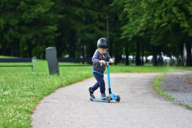 Miğferli genç, parkta mavi scooter kullanıyor. Odaklanmış ve kararlı ifade aktif oyun sırasında konsantrasyonu gösterir. Çevresi yeşillik ve yaz atmosferiyle çevrili..