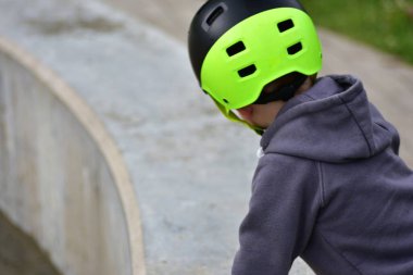 Açık havada oynarken neon yeşil kasklı ve gri kapüşonlu bir çocuk yakın çekimde görülüyor. Park keşfi sırasında kask tasarımı ve çocuk merakına odaklan.