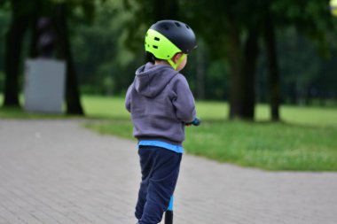 Güvenlik kaskı ve kapüşonlu küçük bir çocuk park yolunda mavi bir scooter sürüyor. Arka plan etkin çocuğu açık havada eğlenirken, yeşil ortamda dengelenirken ve süzülürken yakalar..