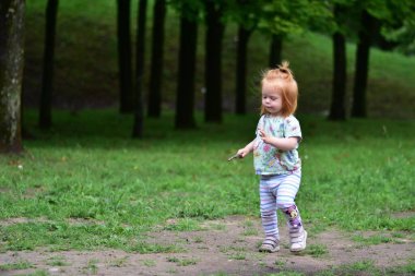 Kızıl saçlı çocuk elinde küçük bir sopayla oyun parkının yanındaki çimenlik yolda yürüyor..