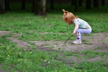 Kırmızı saçlı, tek boynuzlu at tişörtlü, çizgili taytlı çimenli zeminde çömelmiş, dışarıda parkta oynarken sopayla toprağı çiziyor..
