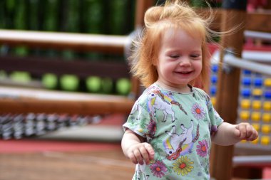 Tek boynuzlu at tişörtü ve çizgili taytlarla gülen kızıl saçlı çocuk parktaki ahşap oyun parkında koşuyor. Neşeli çocukluk ve aktif açık hava oyunu.