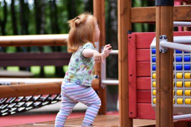 Tek boynuzlu at tişörtü ve çizgili taytlarıyla gülümseyen kızıl saçlı çocuk parktaki ahşap oyun alanında koşuyor. Neşeli çocukluk ve aktif açık hava oyunu.