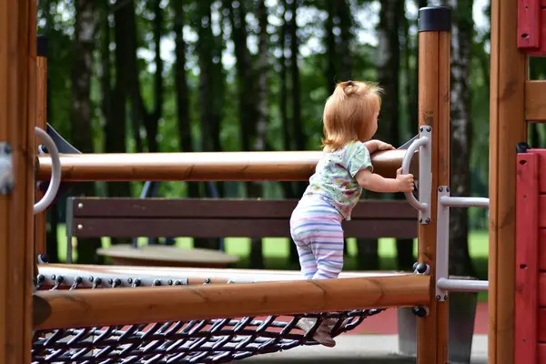 Tek boynuzlu at tişörtü ve çizgili taytlı kızıl saçlı çocuk güneşli bir günde parktaki ahşap oyun parkına tırmanıyor. Açık hava aktivitesi ve çocukluk eğlencesi.