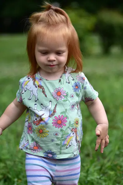 Tek boynuzlu at tişörtü ve çizgili taytlı kızıl saçlı çocuk güneşli bir park gününde oyun parkının yanındaki çimenli alanda yürüyor..