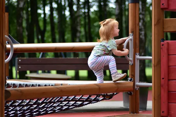 Tek boynuzlu at tişörtü ve çizgili taytlı kızıl saçlı çocuk güneşli bir günde parktaki ahşap oyun parkına tırmanıyor. Açık hava aktivitesi ve çocukluk eğlencesi.