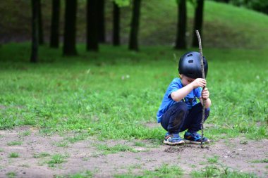 Mavi gömlekli ve siyah kasklı genç bir çocuk parkta yere çömelmiş, iki elinde sopayla çamurda oynuyor. Arka planda otlar ve ağaçlar açık havada barış ortamı oluşturur..