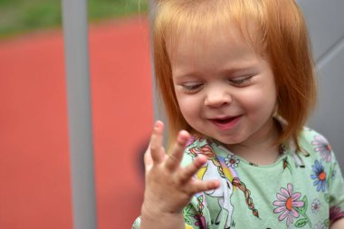 Sevimli kızıl saçlı çocuk merakla gülümsüyor ve eline bakıyor. Tek boynuzlu at ve çiçek desenli yeşil bir gömlek giyiyor. Parlak bir arka planda dışarıda duruyor..