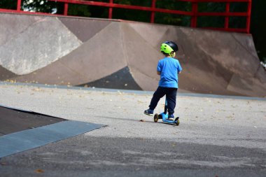 Mavi tişörtlü ve kasklı çocuk kameradan kaykay rampalarına doğru scooter sürüyor. Sahne, hareket ve macera duygusunu yakalar. Küçük çocuk kaykay parkında güvenlik teçhizatıyla scooter sürüyor..