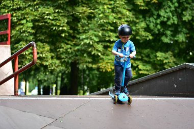 Kasklı genç kaykay rampasında mavi scooter sürüyor, aşağıya doğru ilerlerken odaklanmış görünüyor. Arkaplanı yemyeşil ağaçlar dolduruyor. Mutlu çocuk kaykay parkı rampalarında scooter sürüyor..