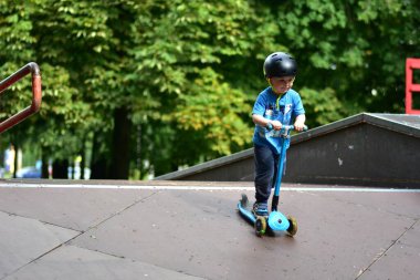 Kasklı genç kaykay rampasında mavi scooter sürüyor, aşağıya doğru ilerlerken odaklanmış görünüyor. Arkaplanı yemyeşil ağaçlar dolduruyor. Mutlu çocuk kaykay parkı rampalarında scooter sürüyor..