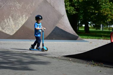 Kasklı genç kaykay rampasında mavi scooter sürüyor, aşağıya doğru ilerlerken odaklanmış görünüyor. Arkaplanı yemyeşil ağaçlar dolduruyor. Mutlu çocuk kaykay parkı rampalarında scooter sürüyor..