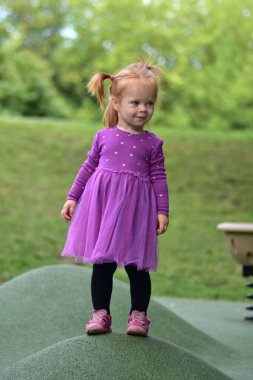 Gülümseyen kızıl saçlı küçük kız mor benekli elbise ve pembe sandaletler bej renkli bahar oyuncağının yanında yeşil çocuk parkında duruyor, yemyeşil arka planı olan güneşli park.